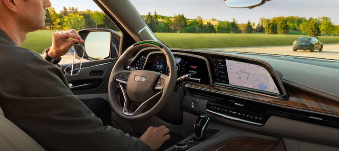 Man wearing gray sweater holding a sungalsses while inside a Cadillac car with refreshing view of nature infront of him