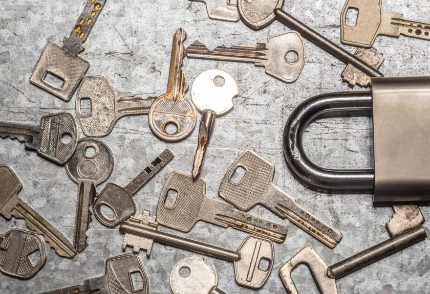 A collection of various metal keys scattered on a textured surface beside a large padlock highlights the importance of lock changes. The diverse shapes and sizes suggest different locks, while the partially visible padlock with its shiny black shackle stands ready for security upgrades.