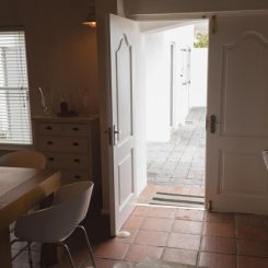 A cozy dining area with a wooden table and white chairs next to a small dresser. The open double doors, recently rekeyed for added security, lead to a patio with tiled flooring. Natural light streams in through a window with blinds, creating a warm and inviting atmosphere.