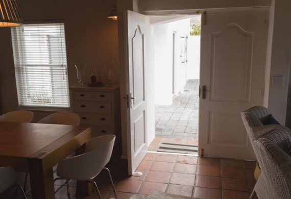 A cozy dining area with a wooden table and white chairs next to a small dresser. The open double doors, recently rekeyed for added security, lead to a patio with tiled flooring. Natural light streams in through a window with blinds, creating a warm and inviting atmosphere.