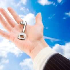 A hand in a business suit holds a vintage key—reminiscent of the Master Key System—against a bright blue sky with scattered clouds. The image evokes themes of opportunity and unlocking potential.