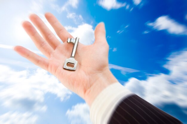 A hand in a business suit holds a vintage key—reminiscent of the Master Key System—against a bright blue sky with scattered clouds. The image evokes themes of opportunity and unlocking potential.