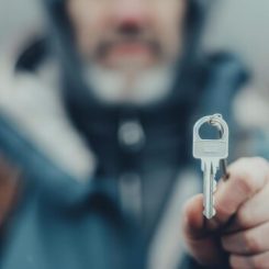 A person in a hooded jacket holds a key towards the camera, blending seamlessly with Locksmith Tips. The background is blurred, emphasizing the key as the focal point of this intriguing image.