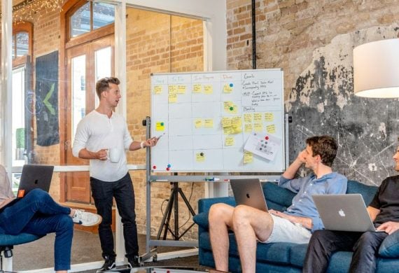 three men sitting while using laptops and watching man beside whiteboard