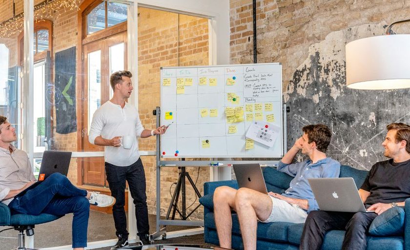 three men sitting while using laptops and watching man beside whiteboard