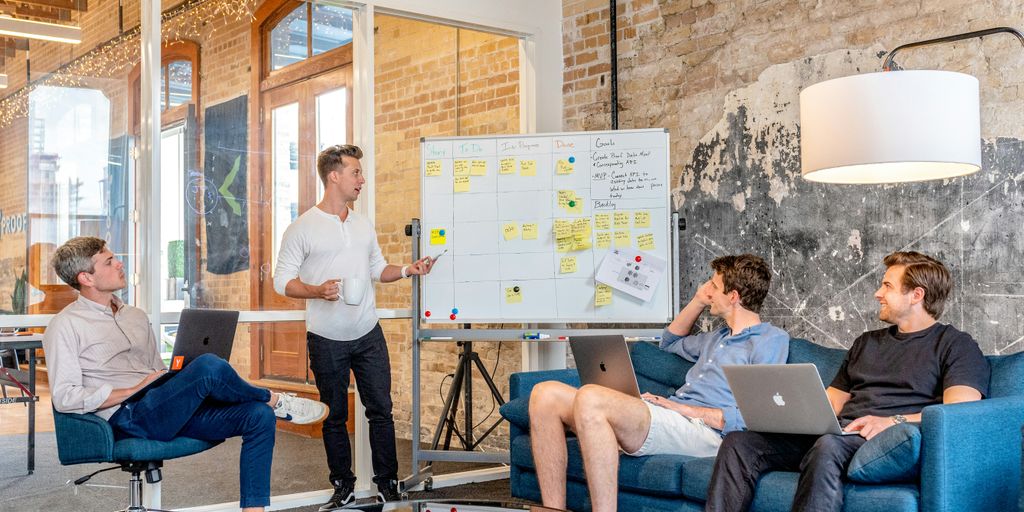 three men sitting while using laptops and watching man beside whiteboard